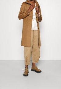 Camelfrakke med en struktureret krave og frontknapper, parret med en cremet top, beige vide bukser og brune snørebåndssko.