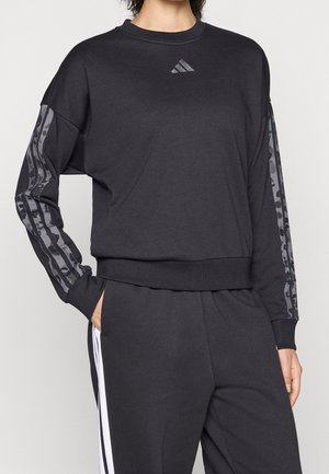Femme portant un sweat-shirt Adidas noir avec une bande à motifs sur les manches et un pantalon noir assorti avec des bandes blanches sur les côtés.