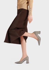 Scarpe con tacco alto in pelle verniciata grigia, con punta affusolata e tacco spesso, abbinate a una gonna marrone, che mostrano un design elegante e una forma moderna.