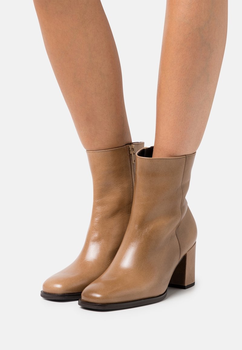 Bottes en cuir beige mi-mollet avec talons carrés, portées sur jambes nues, dotées de fermetures éclair latérales et d'une finition lisse et brillante.
