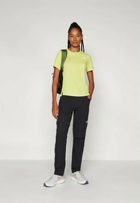 Γυναίκα με πλεξούδες φοράει ένα λαχανί κοντομάνικο μπλουζάκι, μαύρο παντελόνι, λευκά αθλητικά παπούτσια και μια πράσινη τσάντα πλάτης στον έναν ώμο.