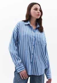 Camicia a righe azzurro chiaro con strisce verticali bianche e rosa, chiusura con bottoni, colletto, taschino sul petto e vestibilità ampia. Il tessuto appare liscio.