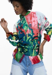 Suéter colorido con patrones mixtos y cuello redondo, que presenta verdes, azules y rojos vibrantes; combinado con una camisa con cuello a contraste.