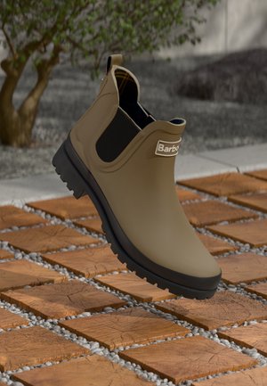 Bottine en caoutchouc marron avec un panneau latéral élastique noir, semelle texturée et logo "Barbour", posée sur une surface en pierre à motif.