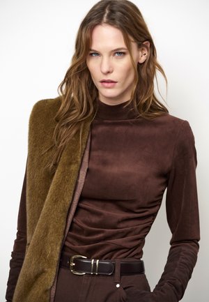 Vrouw met lang bruin haar, die een aansluitende bruine coltrui en riem draagt, met een gestructureerde bruine jas over één schouder gedrapeerd.