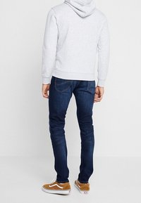 Homme portant un sweat à capuche gris clair, un jean slim bleu foncé et des baskets marron, debout de dos sur fond blanc uni.