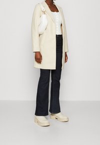 Crème wollen jas, wit geknoopte top, donkerblauwe flared jeans en beige plateau schoenen. Witte handtas met gebogen ontwerp en gouden accenten.