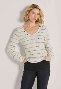 Blonde Frau mit kurzen Haaren, die einen langärmeligen weißen Pullover mit schwarzen horizontalen Streifen und schwarze Hosen trägt, mit den Händen in den Taschen, vor einem neutralen Hintergrund.