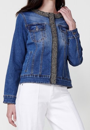 Femme portant une veste en jean bleue avec une bordure décorative en perles le long du col et de l’avant, associée à un pantalon blanc.
