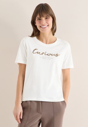 Femme souriante portant un T-shirt blanc avec le texte "Curious" et un pantalon marron, debout contre un mur beige, les mains dans les poches.