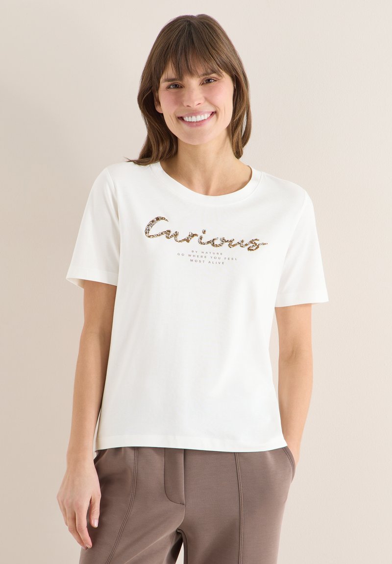 Femme souriante portant un T-shirt blanc avec le texte "Curious" et un pantalon marron, debout contre un mur beige, les mains dans les poches.