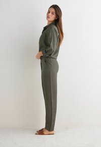 Olivgrön långärmad skjorta och matchande byxor med fickor, gjorda av mjukt tyg. Bruna sandaler med öppen tå fulländar outfiten.