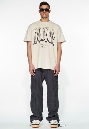 Cream-colored t-shirt with bold black graphic text, paired with dark cargo pants featuring multiple pockets and a relaxed fit.