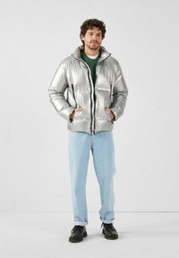 Giacca a sacco argento con finitura lucida, zip frontale, due tasche laterali, cappuccio, indossata sopra un maglione verde e jeans azzurri chiari.