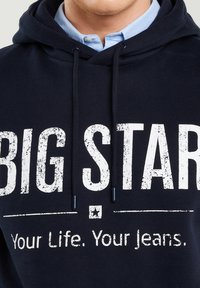 Vyras dėvi tamsiai mėlynį džemperį su gobtuvu ant šviesiai mėlynos marškinių apykaklės, džempyje baltais raidėmis užrašyta: „BIG STAR“ ir „Tavo gyvenimas. Tavo džinsai.“