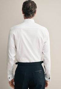 Weißes Hemd mit langen Ärmeln, von hinten gezeigt. Hat einen klassischen Kragen und schwarze Knöpfe an den Manschetten. Kombiniert mit dunklen Hosen.