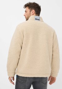 Giacca in pile beige con una texture morbida, colletto alto e etichetta logo sul retro. Presenta polsini elasticizzati e un accento blu sull'orlo.