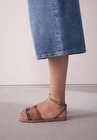 Sandali intrecciati in pelle con una sfumatura marrone e ruggine, cinturino alla caviglia con fibbia e suola piatta, abbinati a una gonna di denim.