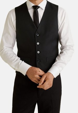 Mann trägt eine schwarze Weste, schwarze Krawatte und weißes Hemd und richtet seine Finger vor einem schlichten weißen Hintergrund.