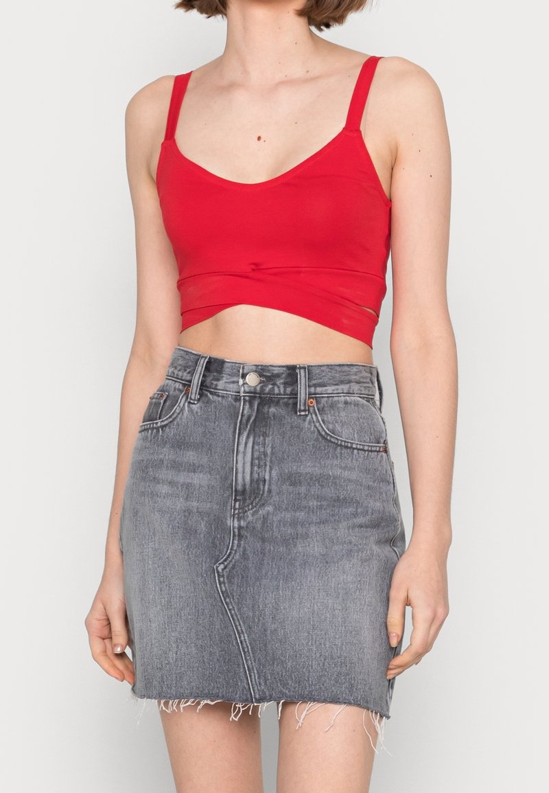 Femme portant un débardeur court rouge et une mini-jupe en jean noir délavé taille haute avec un ourlet effiloché, debout devant un fond uni.