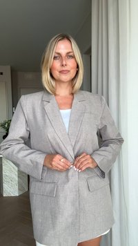 Donna bionda che indossa un blazer oversize grigio chiaro, mentre allaccia il bottone, in piedi all'interno vicino a tende bianche e a un tavolo di marmo.