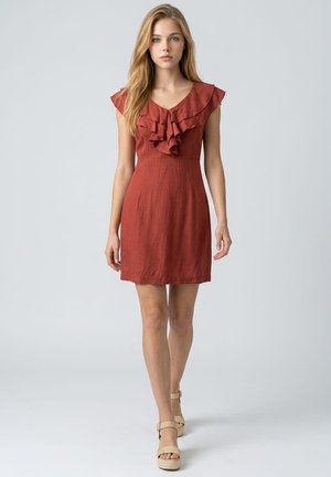 Joven mujer caminando hacia adelante vestida con un vestido sin mangas color óxido con escote fruncido y sandalias beige de plataforma.