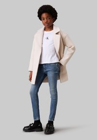 Abrigo beige con solapas anchas sobre una camiseta blanca con el logo de CK, combinado con unos pantalones vaqueros skinny azules y mocasines negros con suela gruesa.