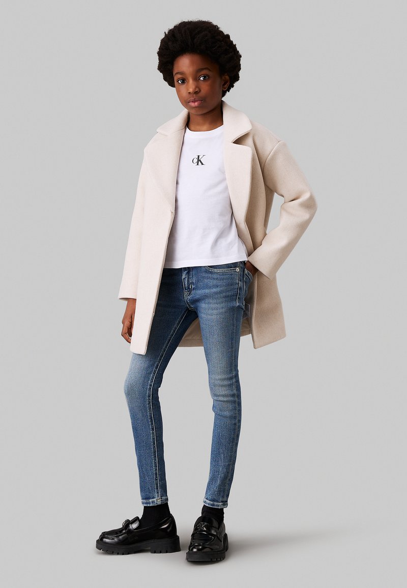 Abrigo beige con solapas anchas sobre una camiseta blanca con el logo de CK, combinado con unos pantalones vaqueros skinny azules y mocasines negros con suela gruesa.
