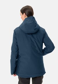 Giacca impermeabile blu scuro con cappuccio, caratterizzata da una texture liscia e polsini regolabili, mostrata di spalle su uno sfondo chiaro.