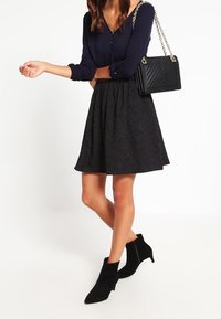 Une femme porte une blouse bleu foncé, une jupe noire texturée, des bottines noires et porte un sac à main matelassé noir avec une chaîne dorée.