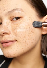 Zbliżenie na twarz o jasnej karnacji i wyraźnych piegach, nakładającej krem pędzlem. Gładka struktura skóry, subtelny kolor ust.