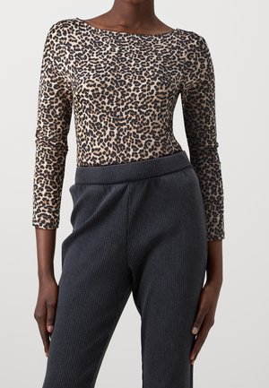 Vrouw draagt een langärmelig top met luipaardprint en hooggetailleerde, donkergrijze ribbroek tegen een effen achtergrond.