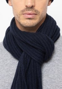 Marineblauwe geribbelde gebreide sjaal van zacht materiaal, om de nek gewikkeld, met een gestructureerde afwerking tegen een grijze trui achtergrond.
