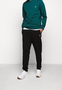 Sweatshirt en coton teal avec col rond, associé à un jogging en coton noir avec un logo brodé. Baskets blanches avec semelles en gomme.