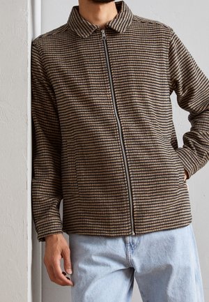 Mand iført brun og sort striped lynlåstrøje med krave og lyseblå jeans, står op ad hvid væg.