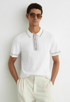 Bărbat purtând un polo alb cu mâneci scurte, cu deschidere cu nasturi și margini bej, pantaloni plisati crem și ochelari de soare cu lentile maronii, cu mâinile în buzunare.