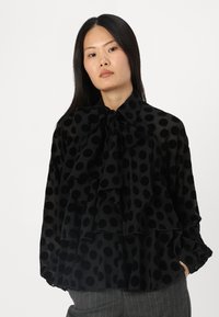 Black sheer blouse with large textured polka dots, bow tie collar, and layered frill design. Long sleeves with gathered cuffs.