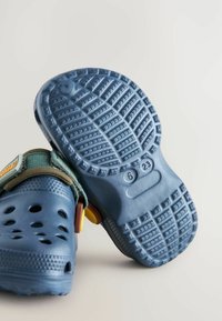 Sabots bleus avec trous de ventilation, semelle en caoutchouc texturée, sangle verte ajustable et détail en jaune. Légers et faciles à porter.