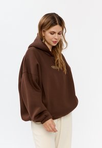 Bruine oversized hoodie met een frontale kangaroo zak en rib gebreide manchetten. Bevat een klein geborduurd logo op de borst. Zachte stof textuur.