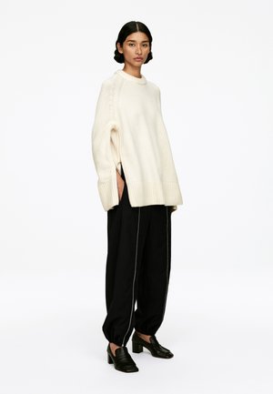 Übergroßer cremefarbener Pullover mit geripptem Saum und seitlichen Schlitzen, kombiniert mit schwarzen, taillierten Hosen mit weißen Paspeln und klobigen schwarzen Schuhen.