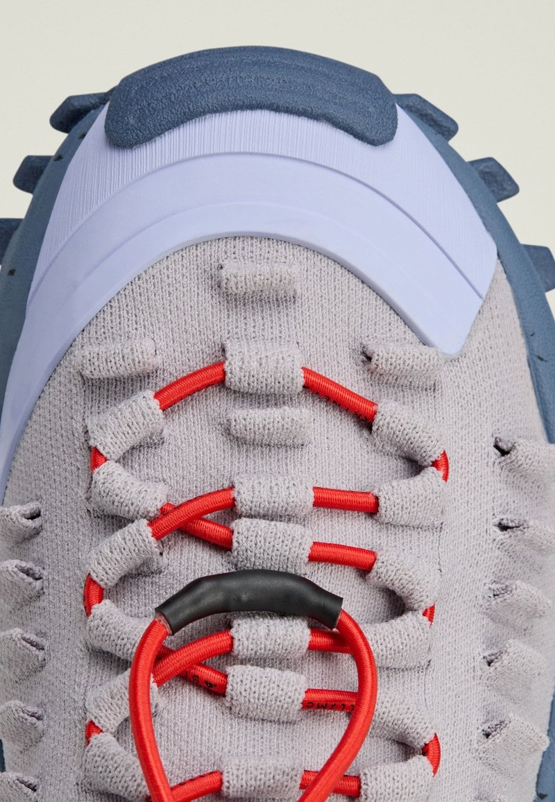 Scarpone sportivo grigio in tessuto con tomaia texturizzata, lacci elastici rossi e dettagli blu. Presenta un collare imbottito e una suola in gomma sagomata.