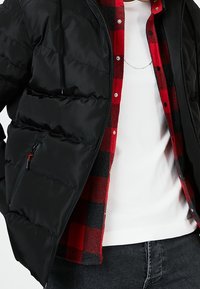 Chaqueta acolchada negra superpuesta sobre una camisa de franela a cuadros roja y gris. Camiseta blanca de cuello redondo visible debajo. Cuenta con un bolsillo con cremallera.