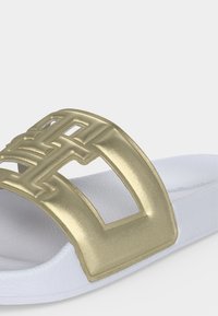 Sandale blanche à enfiler avec semelle épaisse et large bride dorée métallique présentant un motif géométrique découpé sur le pied.