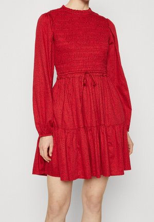 Czerwona sukienka z marszczonym gorsetem, długimi rękawami i wielowarstwową spódnicą. Posiada talię z troczkiem oraz drobny wzór czarnych kropek na całej powierzchni.