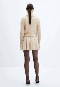 Maglione beige a maglia con una trama a coste, abbinato a una gonna plissè beige. La modella indossa calze nere trasparenti e scarpe con tacco alto.