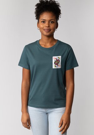 Jeune femme aux cheveux bouclés foncés portant un T-shirt bleu sarcelle avec une carte à jouer Reine de Cœur imprimée sur la poitrine.