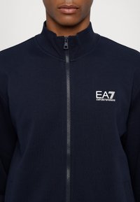Giacca a zip navy in morbido tessuto con colletto alto, caratterizzata da un logo bianco EA7 sul lato sinistro del petto e una cerniera metallica con un tirante.