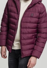 Burgundowa kurtka puchowa z poziomym pikowanym wzorem, z przodem na zamek błyskawiczny, odczepianym kapturem i kieszeniami bocznymi, noszona na kremowym swetrze.