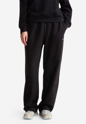Personne portant un pantalon de survêtement noir ample et un sweat-shirt noir avec des baskets beiges et blanches, debout sur un fond blanc.