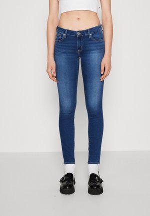 Vrouw draagt mid-rise blauwe skinny jeans, wit cropped topje, witte sokken en zwarte loafers, staand tegen een effen achtergrond.
